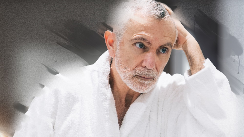 Oudere man in badjas kijkt doordacht in beslagen badkamerspiegel